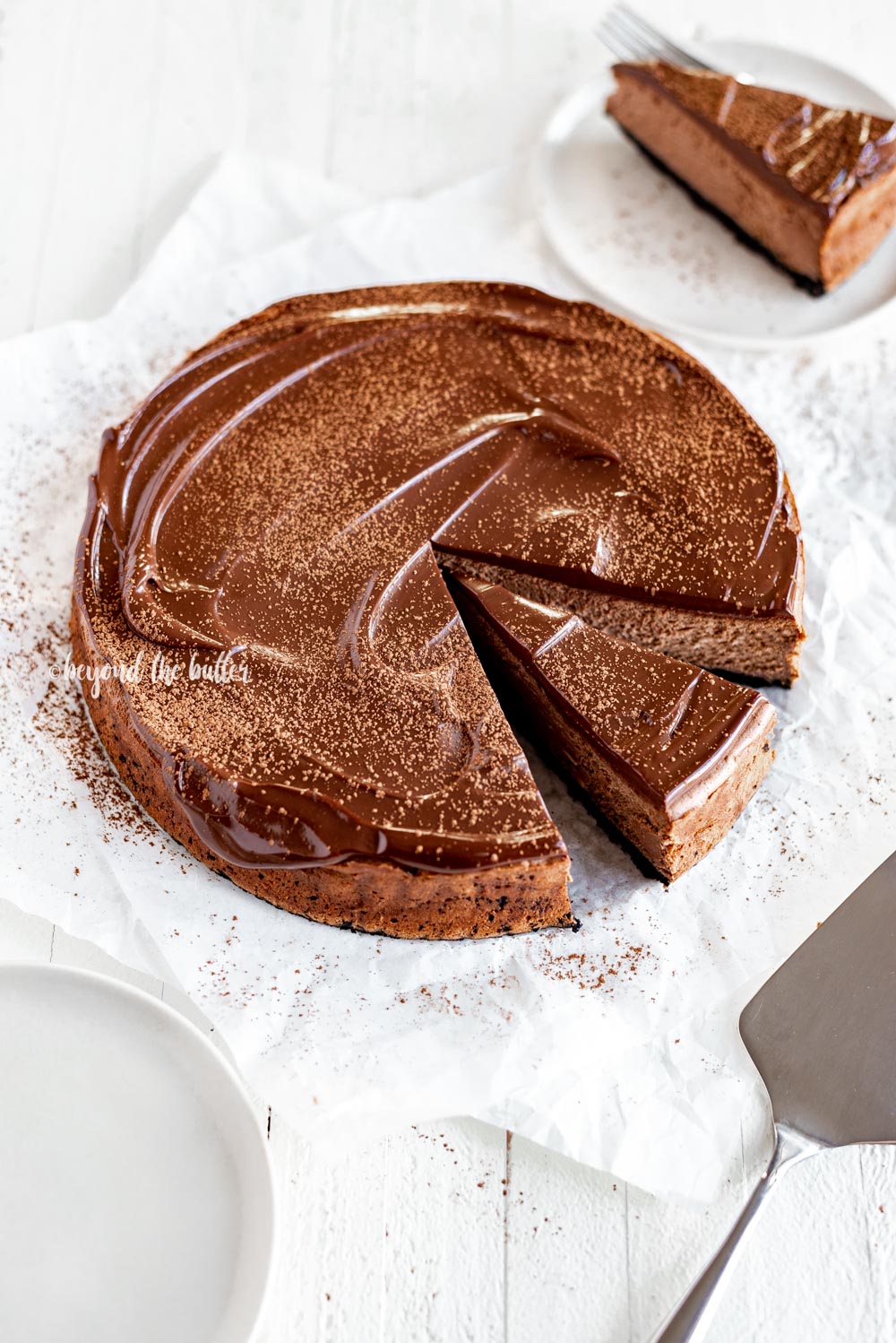 Overhead angled image of Triple Chocolate Mocha Cheesecake dusted with cocoa and a slice removed | All Images © Beyond the Butter, LLC