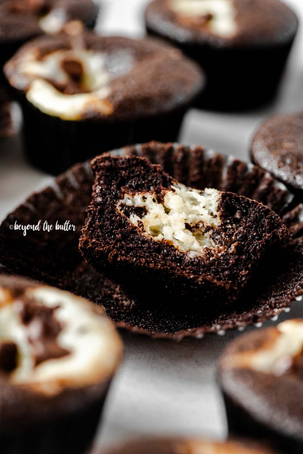Close up angled image of black bottom cupcakes randomly placed with a half sliced cupcake on gray background | All Images © Beyond the Butter™