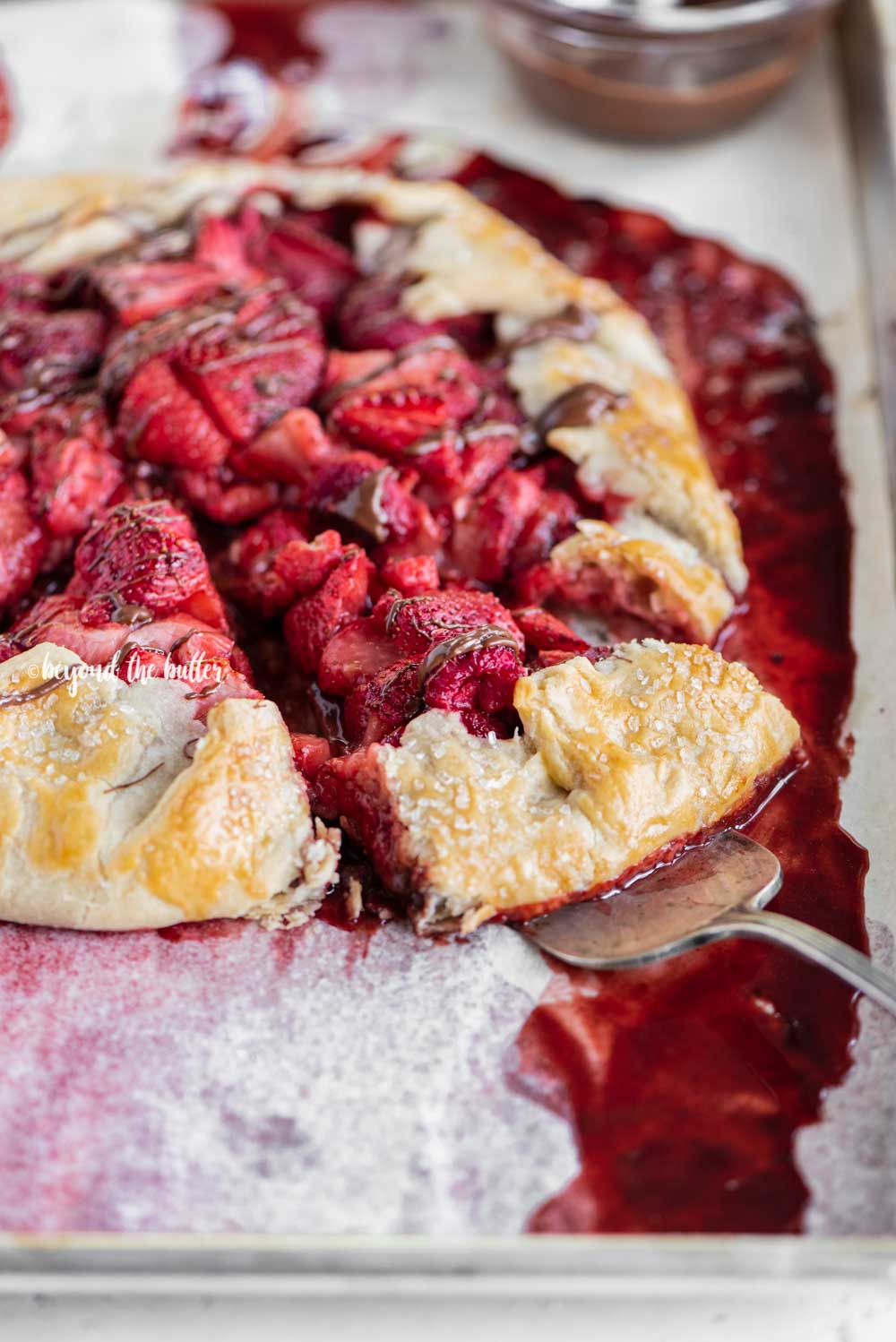 Angled image of a Berry Nutella Galette on a baking sheet with Nutella drizzled over the top and 2 slices cut | All Images © Beyond the Butter™