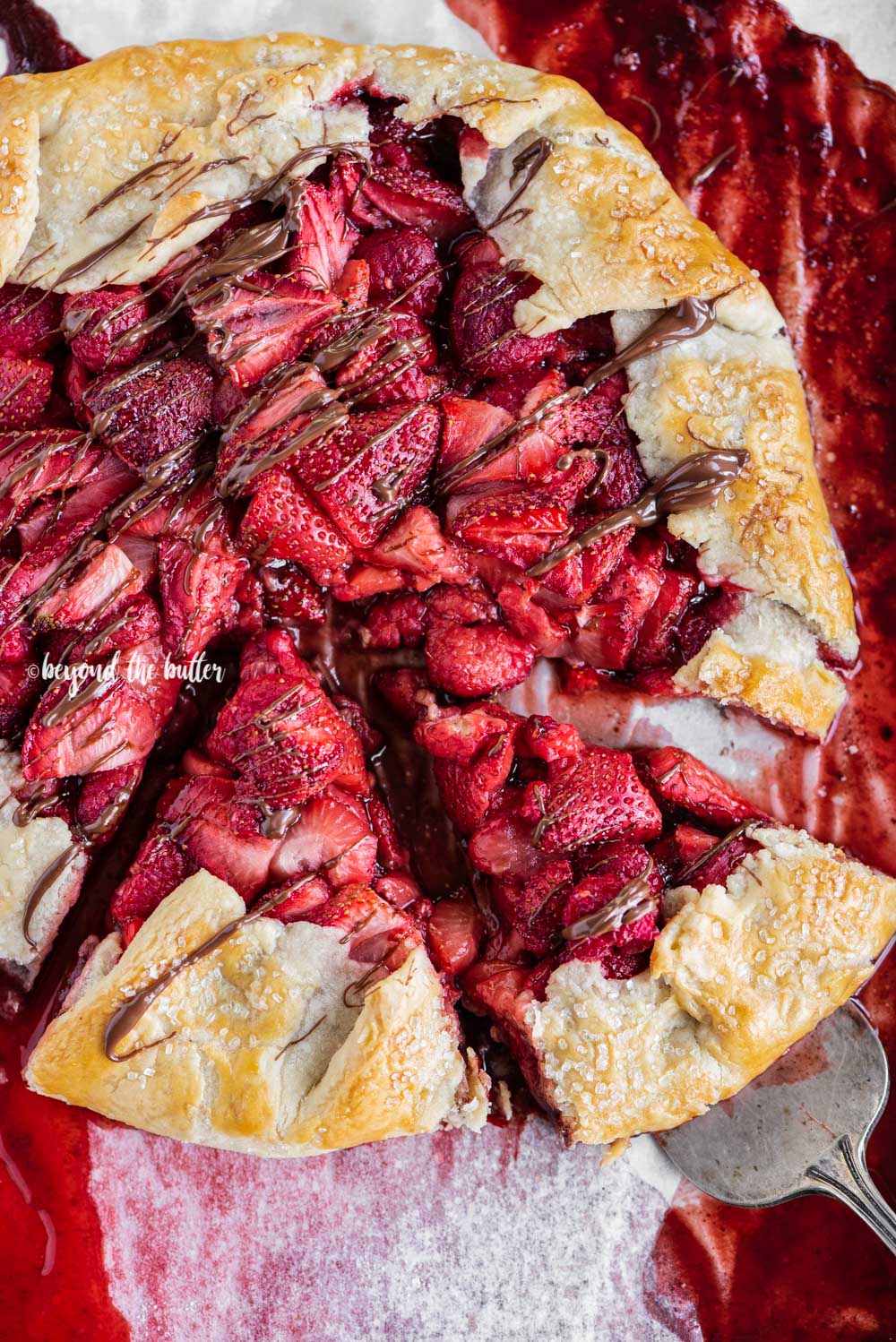 Overhead image of a Berry Nutella Galette on a baking sheet with Nutella drizzled over the top and 2 slices cut | All Images © Beyond the Butter™