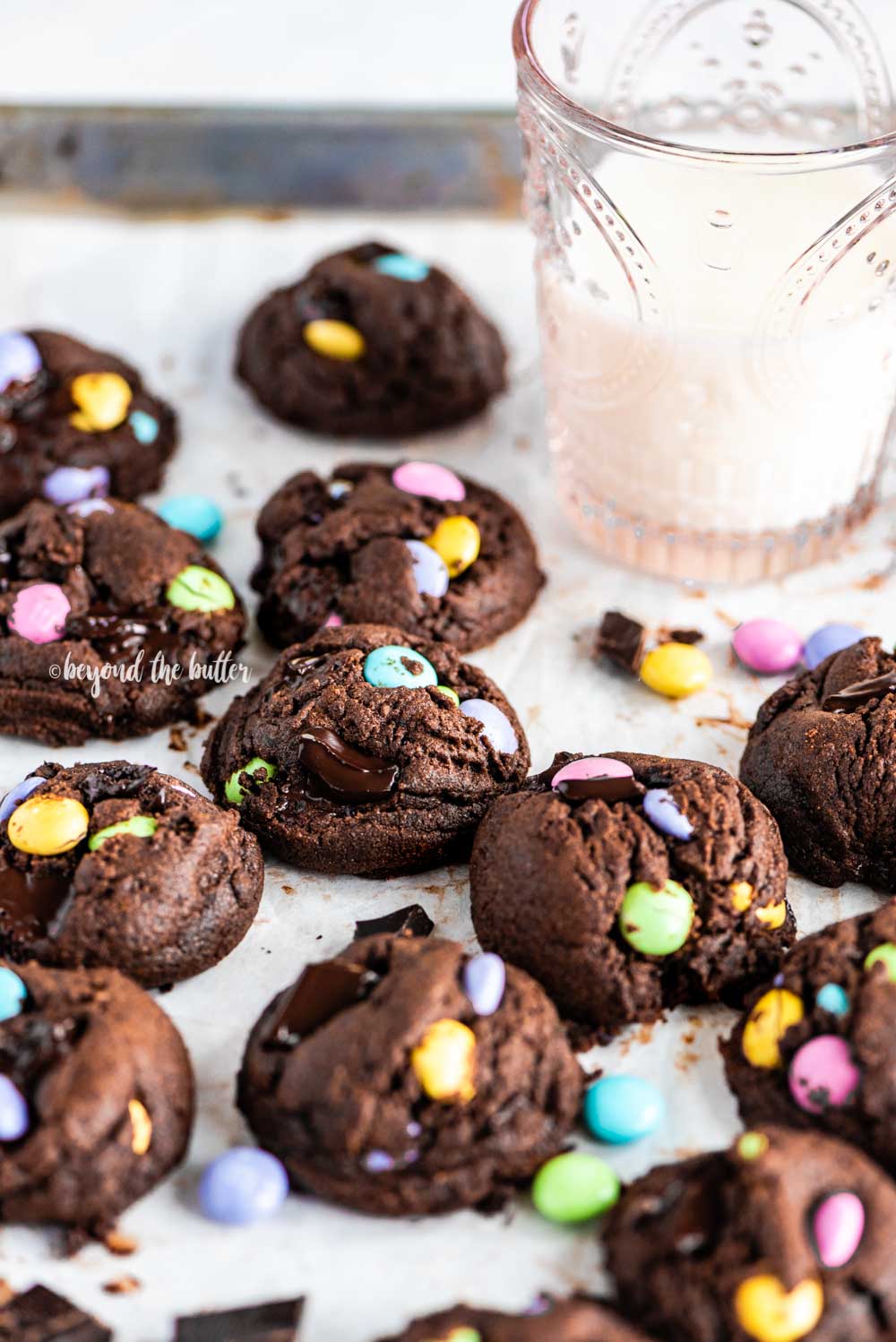 Angled image of just baked double chocolate chunk m&m cookies on a parchment lined baking sheet and a glass of milk above it | All Images © Beyond the Butter™