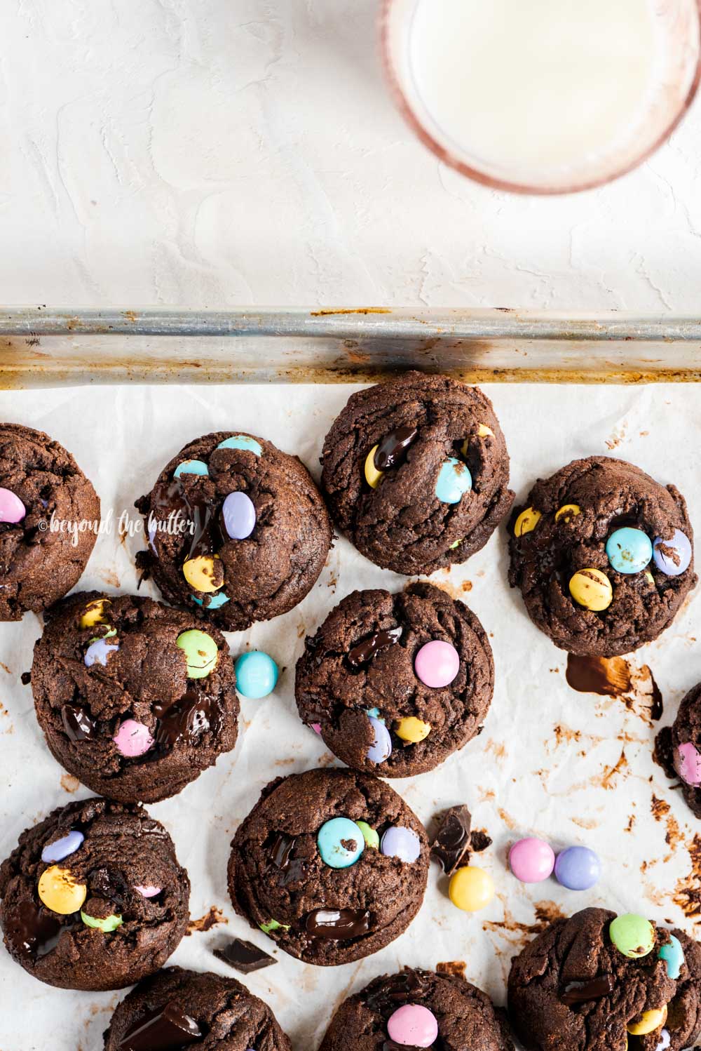 Overhead image of just baked double chocolate chunk m&m cookies on a parchment lined baking sheet and a glass of milk above it | All Images © Beyond the Butter™