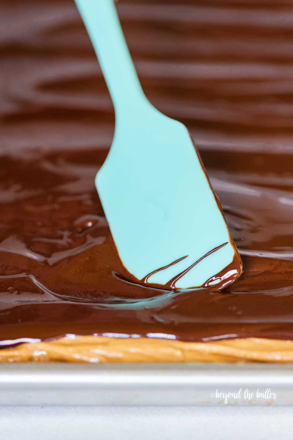 Angled view of blue spatula spreading chocolate over the top of peanut butter tandy kakes | All Images © Beyond the Butter™