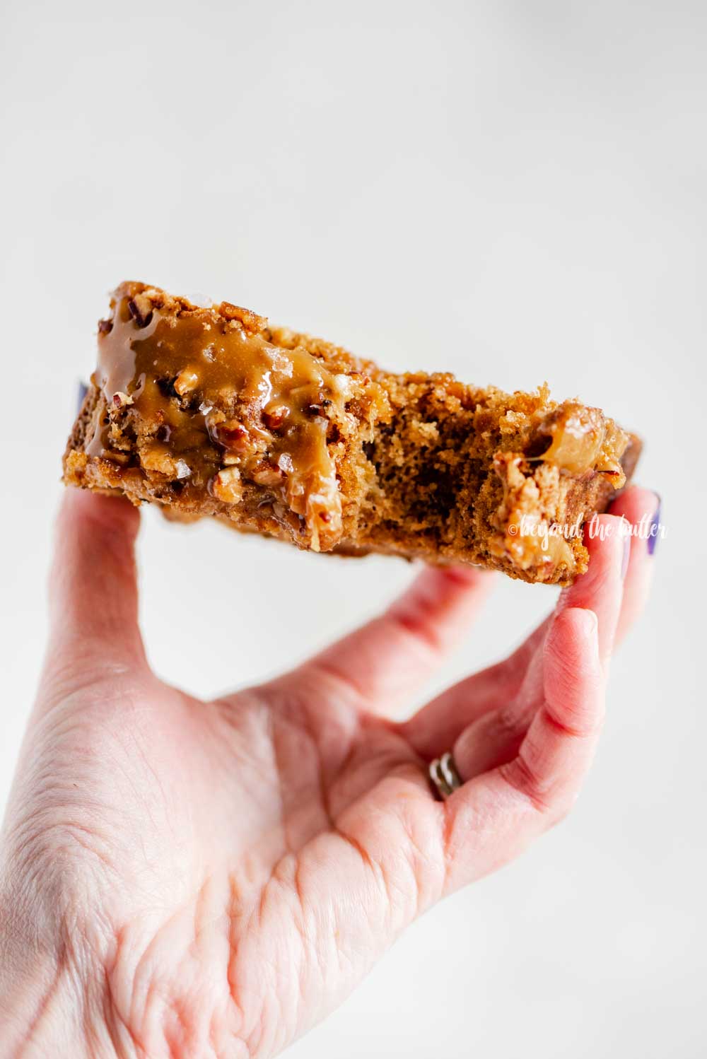 Image of woman's hand hold a slice of salted caramel banana nut bread to show the top with a bite out of it | All Images © Beyond the Butter™