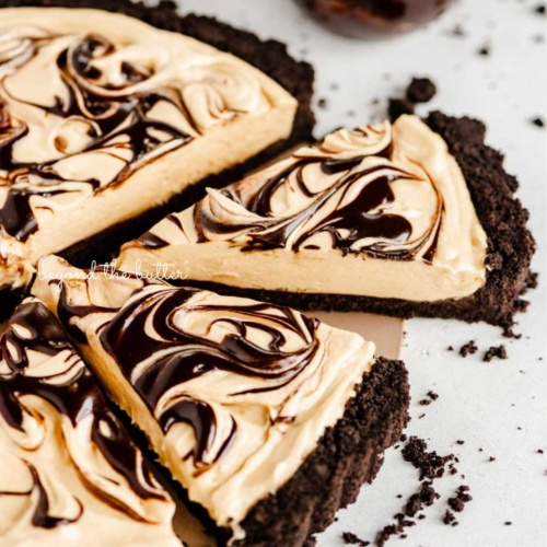 Round chocolate peanut butter tart swirled with chocolate syrup and cut into slices with a bowl of chocolate syrup next to it surround by a few chocolate sandwich cookie crumbs on a white speckled background.