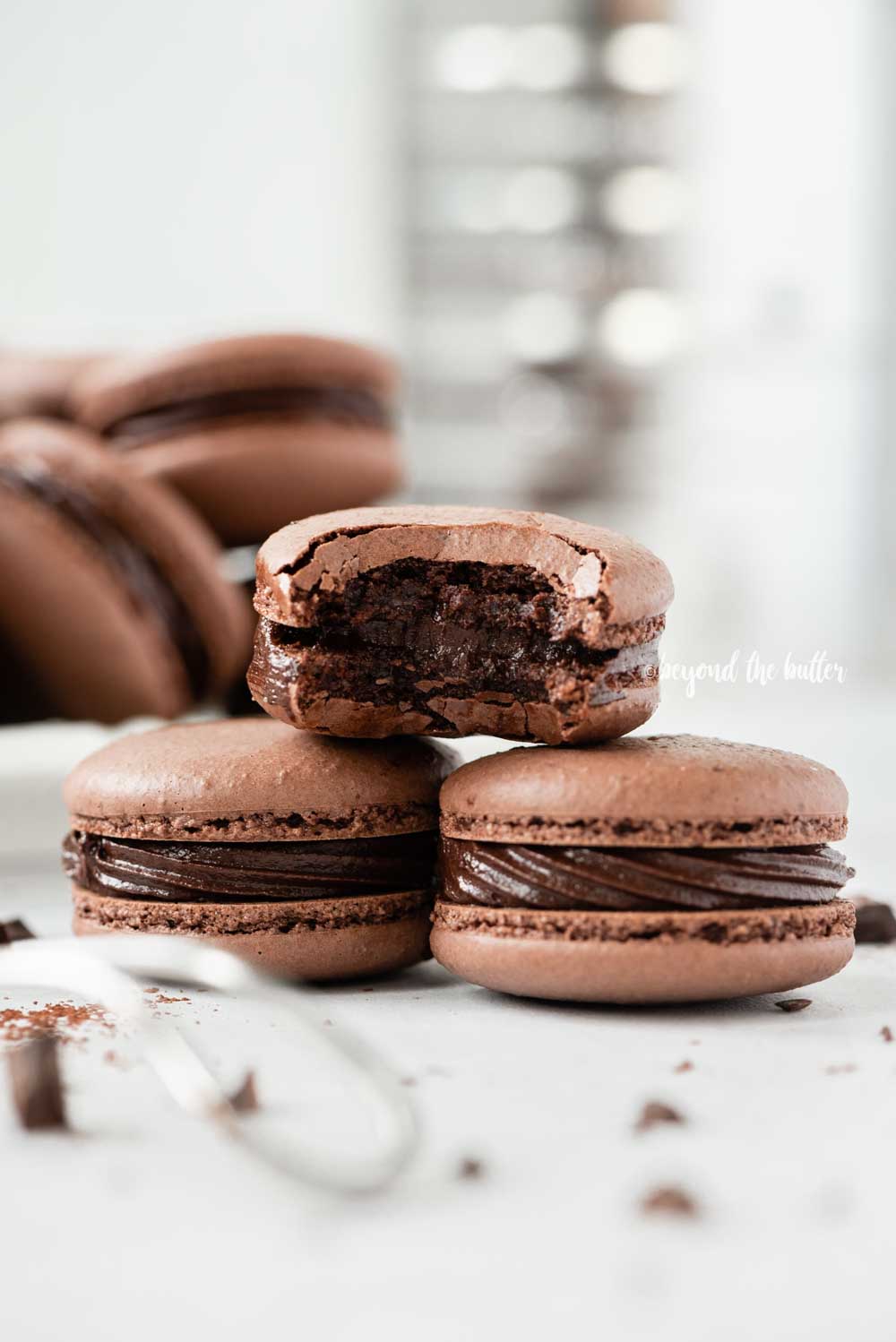Small stack of chocolate macarons with a bite taken out of the top macaron with a bigger stack in the background with a sifter | All Images © Beyond the Butter™
