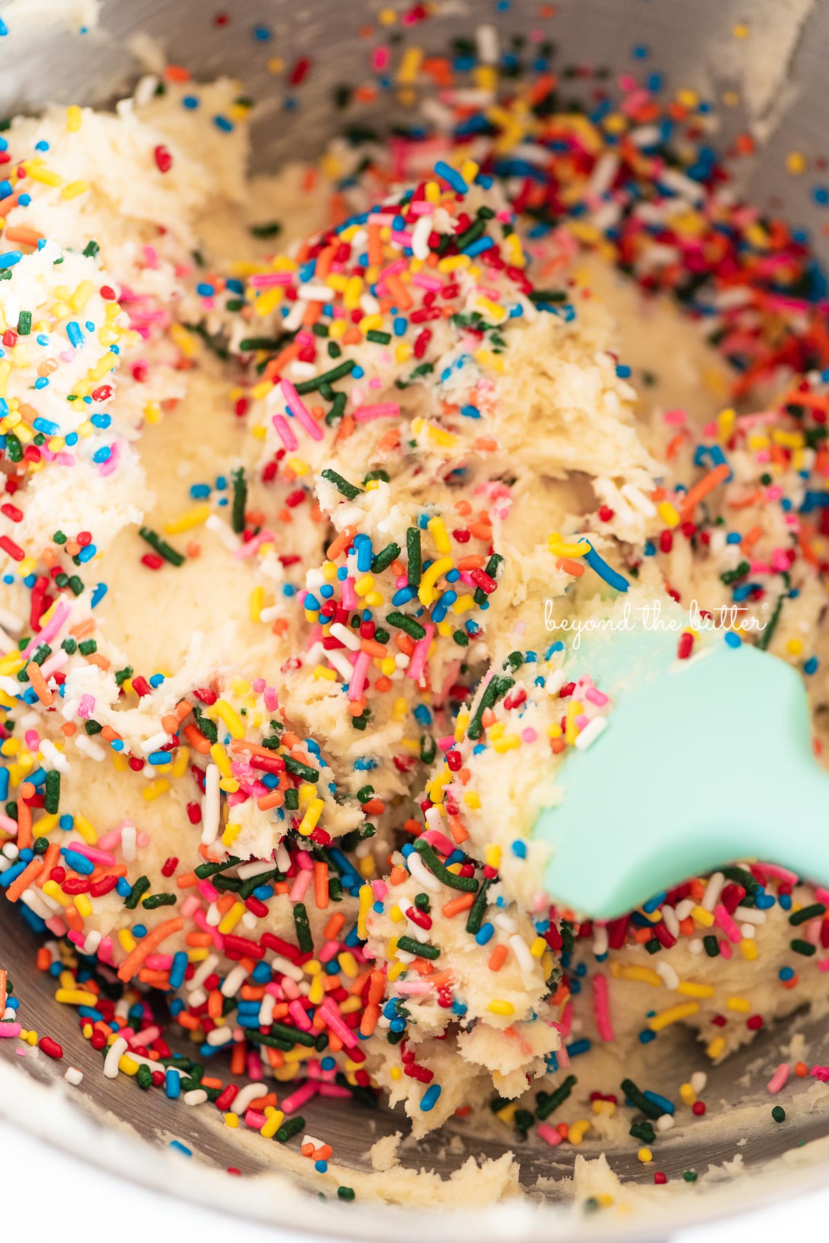 A light teal blue spatula stirring rainbow sprinkles into cookie dough to make funfetti butter cookies.