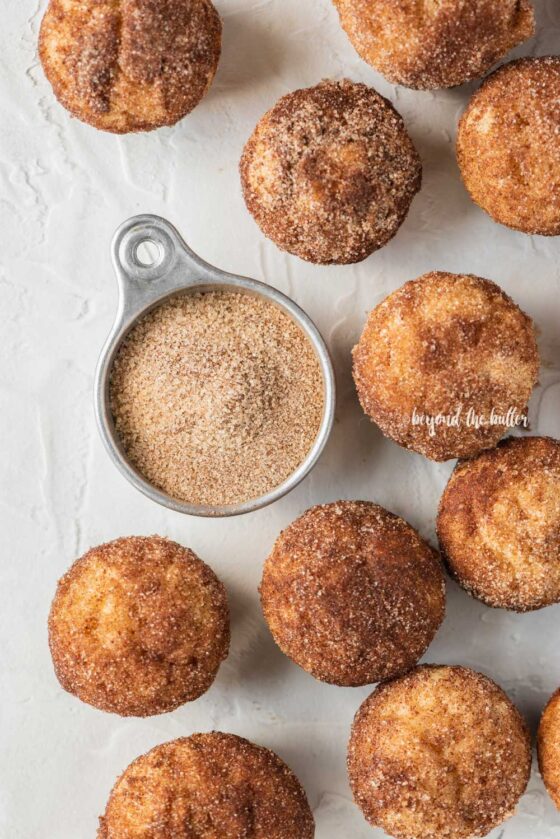 Overhead image of mini french breakfast puffs on a white background with a measuring cup filled with cinnamon sugar mix | All Images © Beyond the Butter™