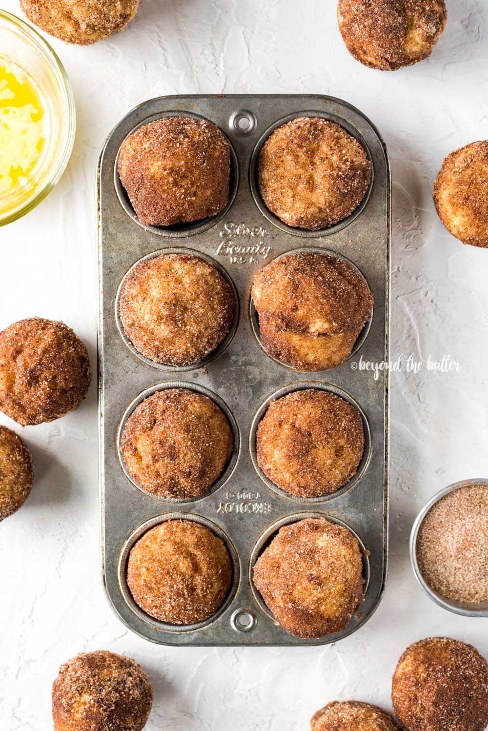 Overhead image of mini french breakfast puffs laid on top of a mini muffin tin with bowls of melted butter and cinnamon sugar around it | All Images © Beyond the Butter™