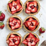 Overhead image of 6 mini strawberry nutella tarts on white stucco background with strawberries and forks around them.