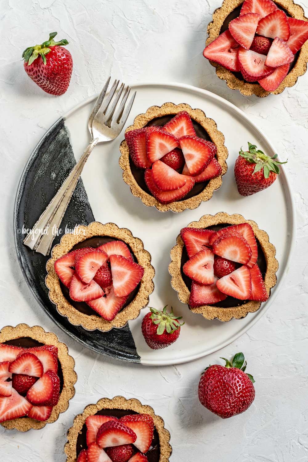 Overhead image of 6 mini strawberry nutella tarts on a black and white serving plate and randomly placed strawberries | All Images © Beyond the Butter™