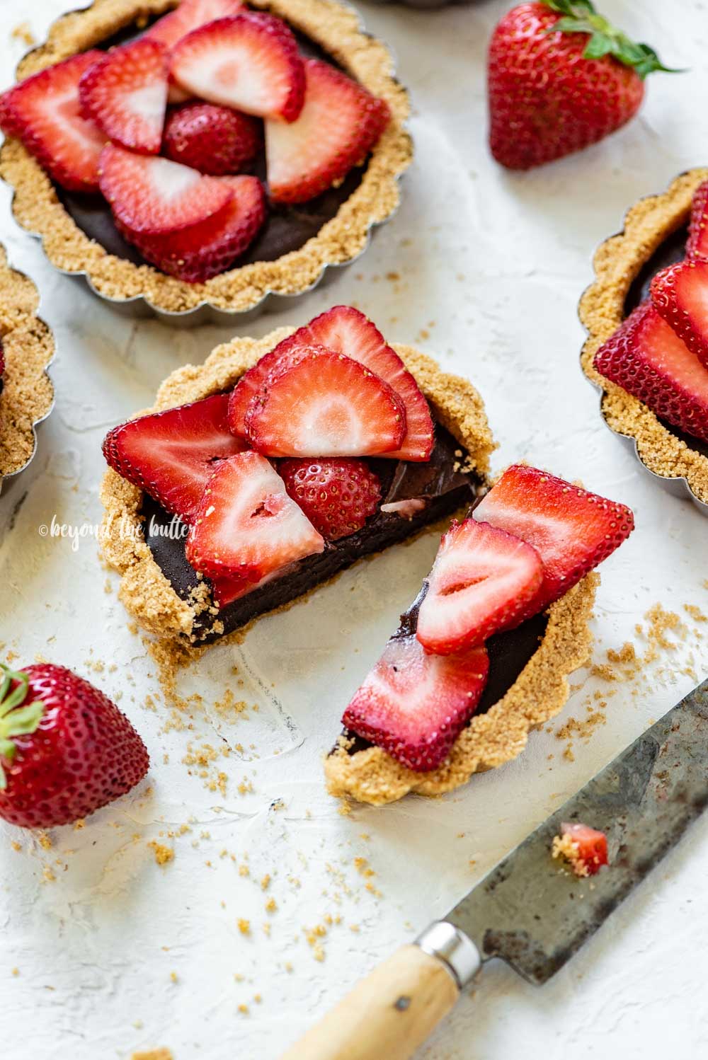 Angled image of mini strawberry nutella tarts on white stucco background with one sliced in half and opened | All Images © Beyond the Butter™