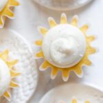 Overhead image of vanilla cupcakes with vanilla buttercream frosting sprinkled with sparkling sugar grouped close together on white background and small ceramic plates.