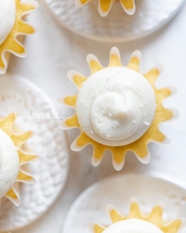 Overhead image of vanilla cupcakes with vanilla buttercream frosting sprinkled with sparkling sugar grouped close together on white background and small ceramic plates.