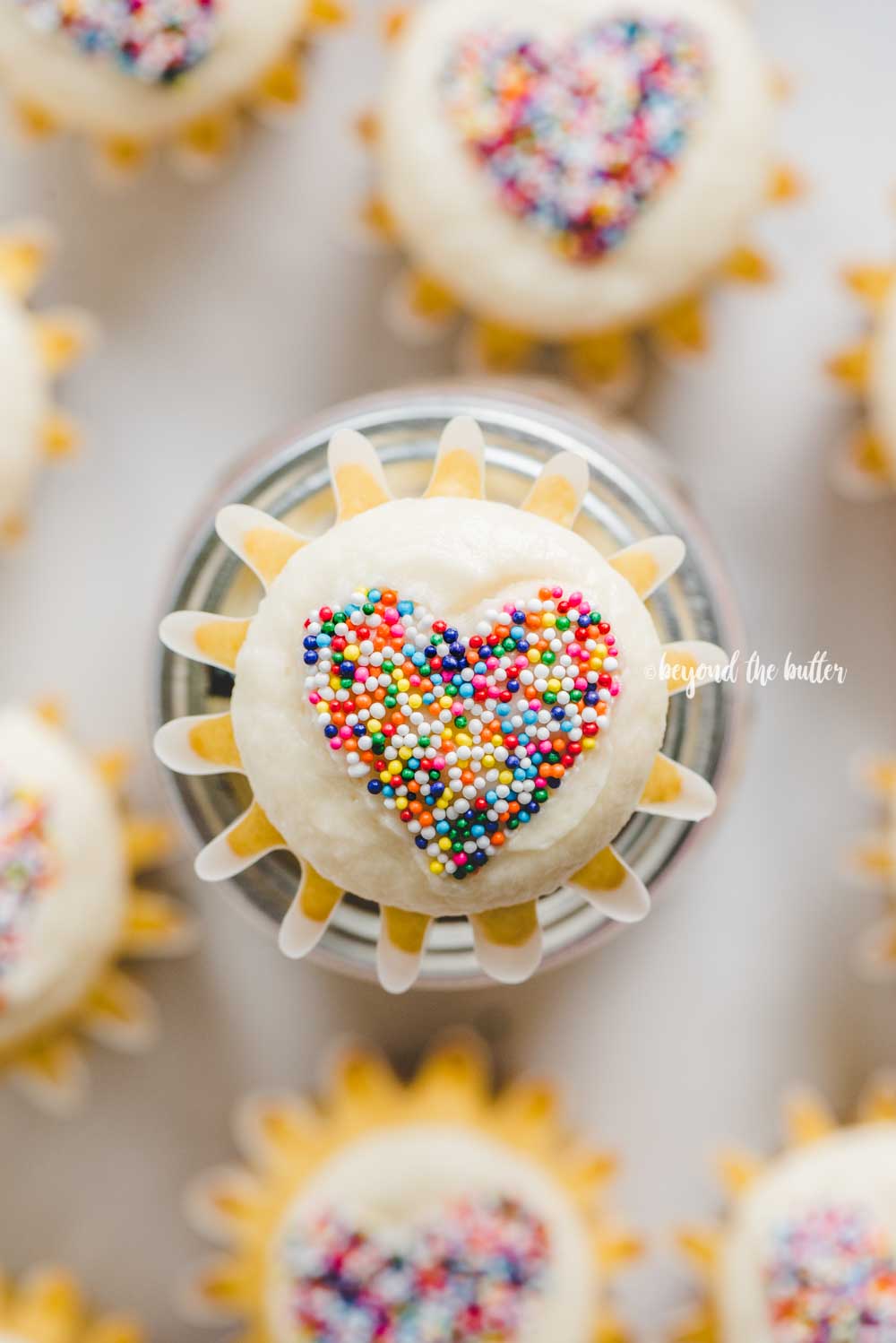 Overhead image of sprinkled heart cupcakes on white background with center cupcake on top of jar.