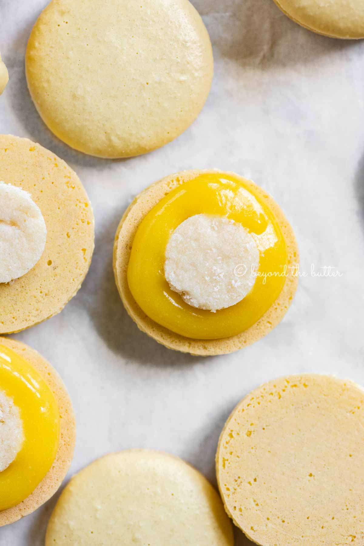 Angled image of lemon bar macarons half opened topped with shortbread crust centers and lemon curd filling | All Images © Beyond the Butter™