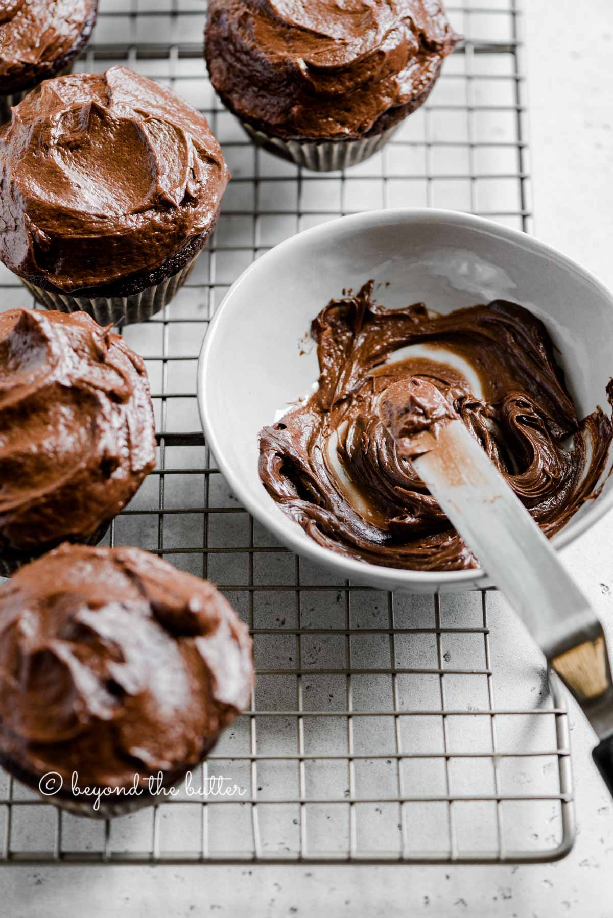Angled image of chocolate cupcakes on cooling rack with small bowl of chocolate frosting and offset spatula | All Images © Beyond the Butter™