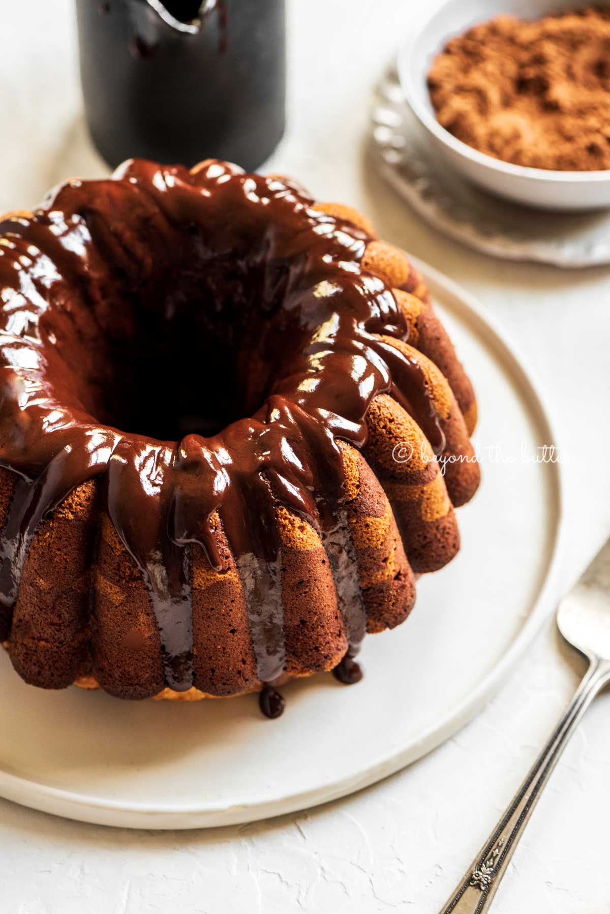 Angled image of chocolate glazed marble bundt cake on black and white cake platter, small bowl of cocoa, and 2 forks | All Images © Beyond the Butter™