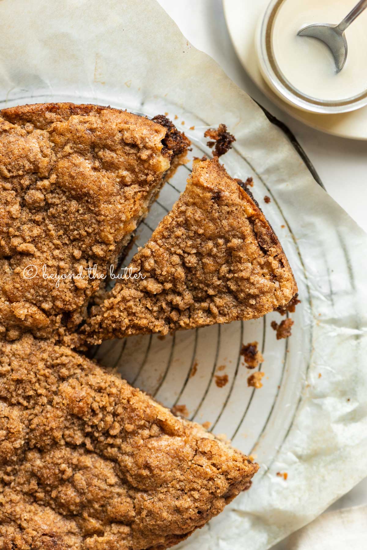 Apple coffee cake on a parchment paper lined wire cooling rack with slices removed and a simple vanilla glaze to the side | All Images © Beyond the Butter™