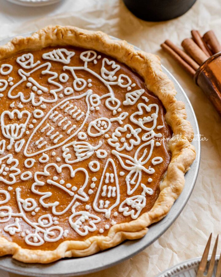 Angled image of pumpkin pie with a piped cream cheese frosting decoration surrounded by forks, cinnamon sticks, plates, and small pitcher of milk.