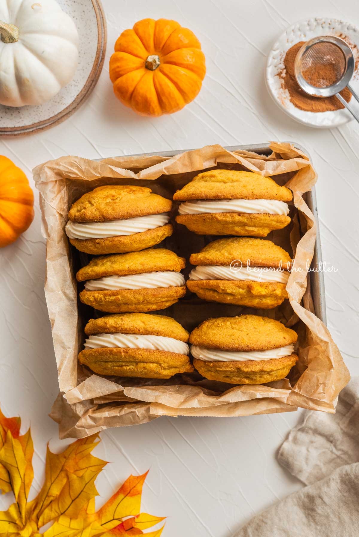 Overhead image of pumpkin whoopie pies in parchment paper lined baking tin on white background with mini pumpkin, fall leaves, dessert plates, and napkin around them | All Images © Beyond the Butter™