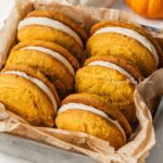 Angled image of pumpkin whoopie pies in parchment paper lined baking tin surrounded by mini orange and white pumpkins on a white background.