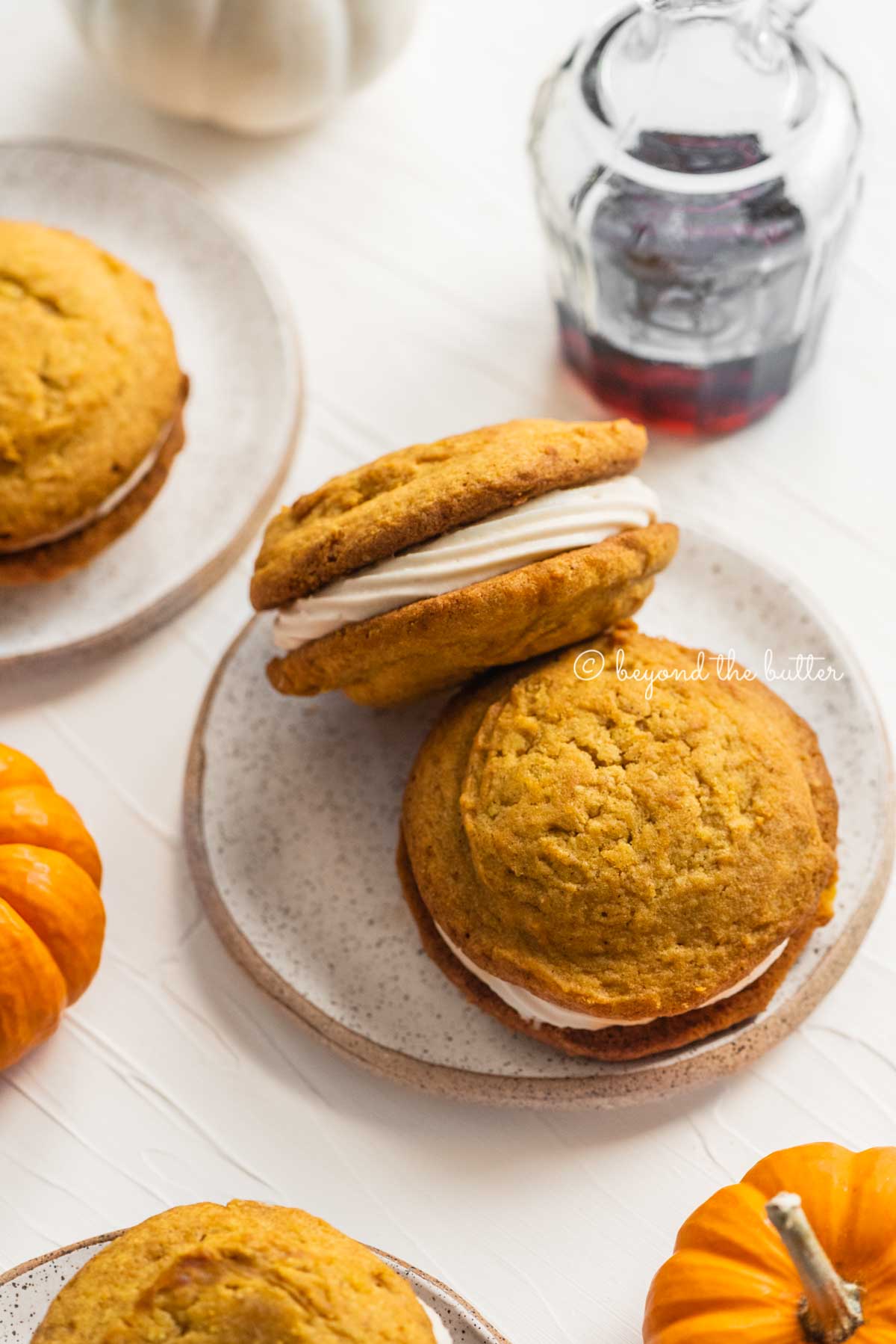 Pumpkin whoopie pies on speckled dessert plates with randomly placed mini pumpkins and a glass of maple syrup | All Images © Beyond the Butter™