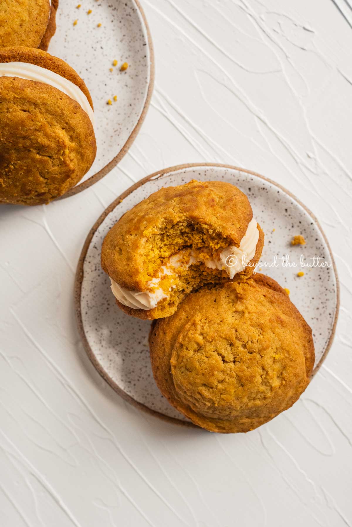 Pumpkin whoopie pies with one half eaten resting on 2 speckled dessert plates | All Images © Beyond the Butter™