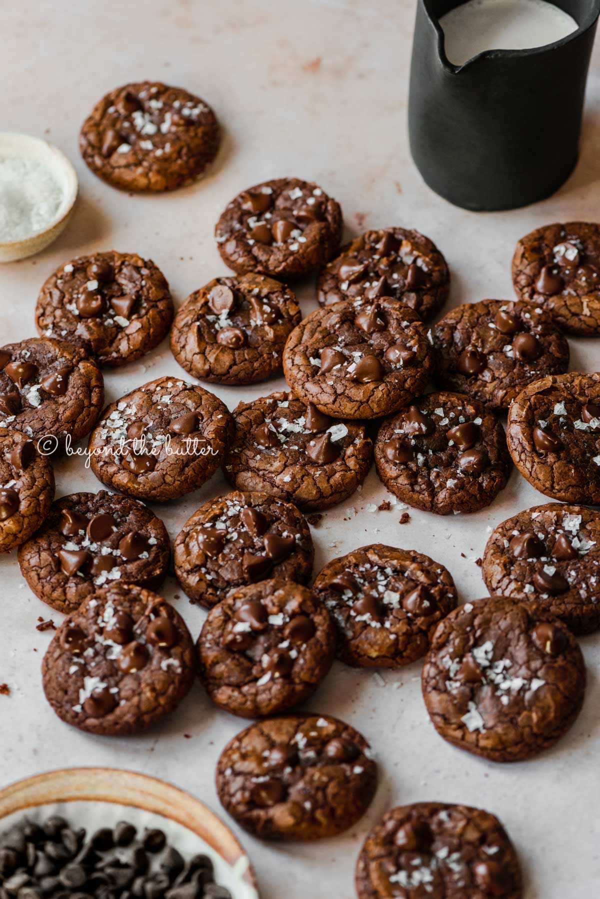 Salted chocolate chip brownie cookies randomly placed on an abstract light brown background with small black pitcher of milk and bowl of sea salt flakes to the side | All Images © Beyond the Butter®