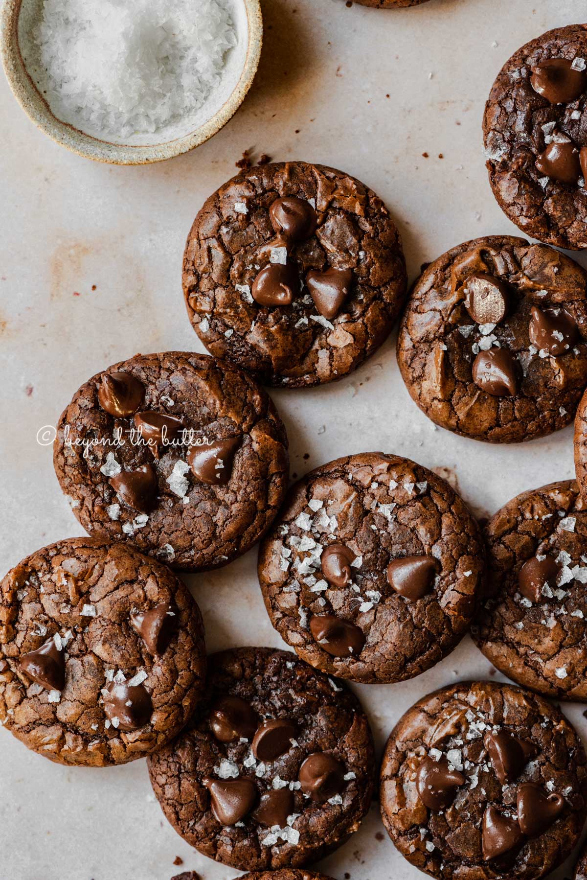 Salted chocolate chip brownie cookies randomly placed on an abstract light brown background with small bowl of sea salt flakes to the side | All Images © Beyond the Butter®