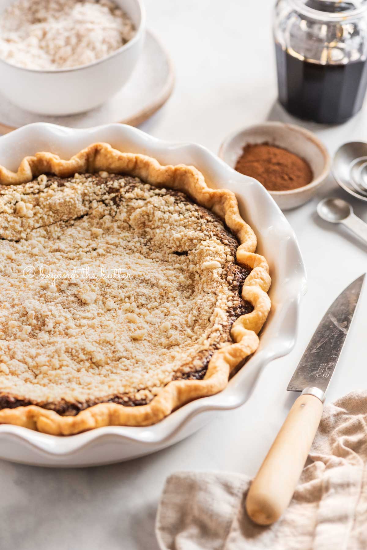 pie plate with uncut shoo fly pie with bowls of crumb topping and cinnamon above and a knife, measuring spoons, and molasses | All Images © Beyond the Butter™
