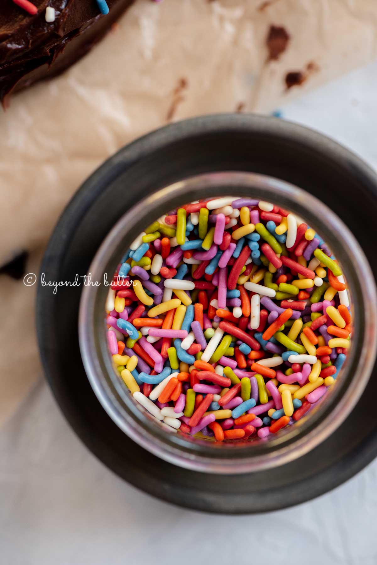 Small bowl of rainbow sprinkles on a dark plate with chocolate cake next to it | All Images © Beyond the Butter®