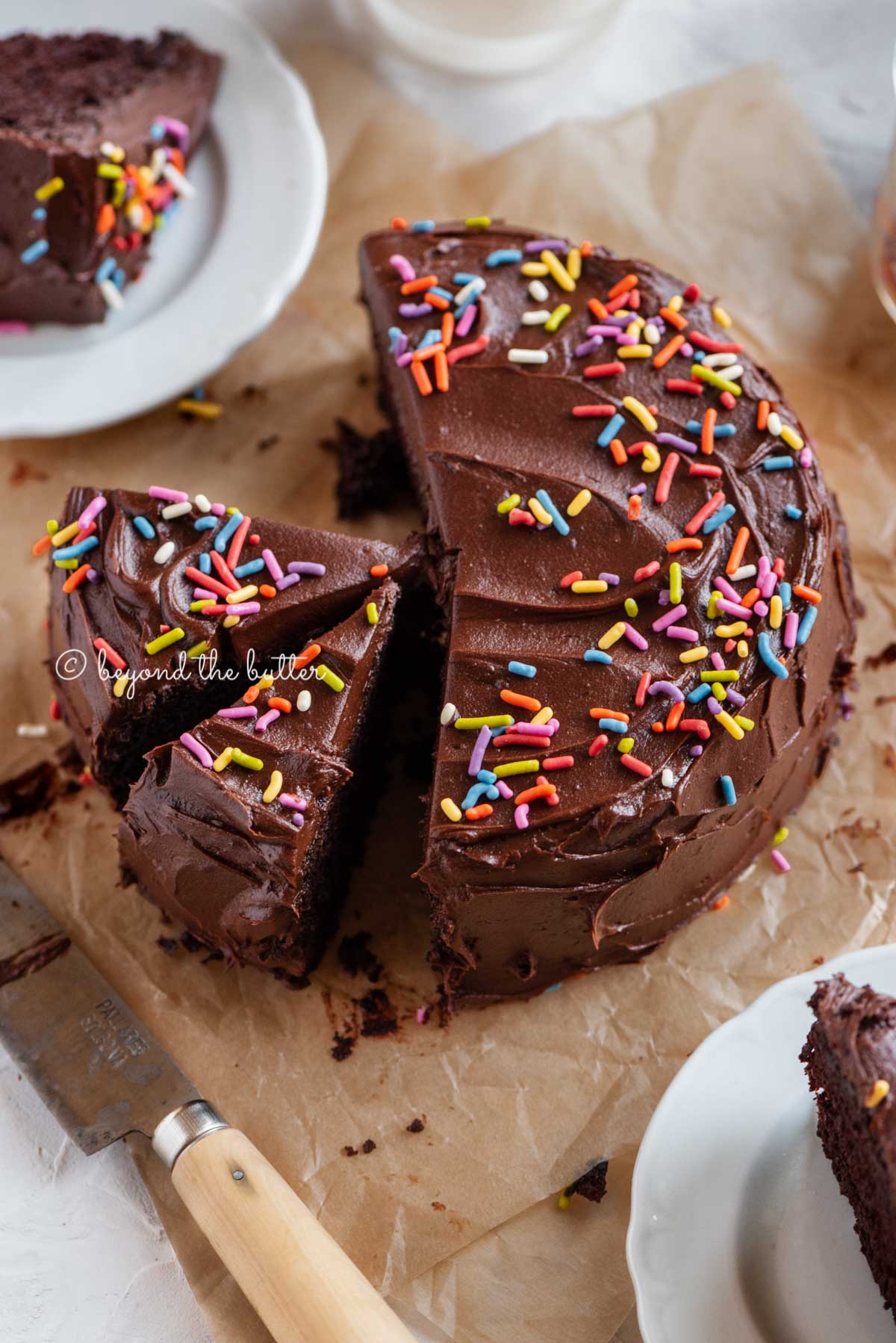 Angled image of sliced single layer chocolate cake on parchment paper with slices of cake on small dessert plates around it and a knife on parchment paper.