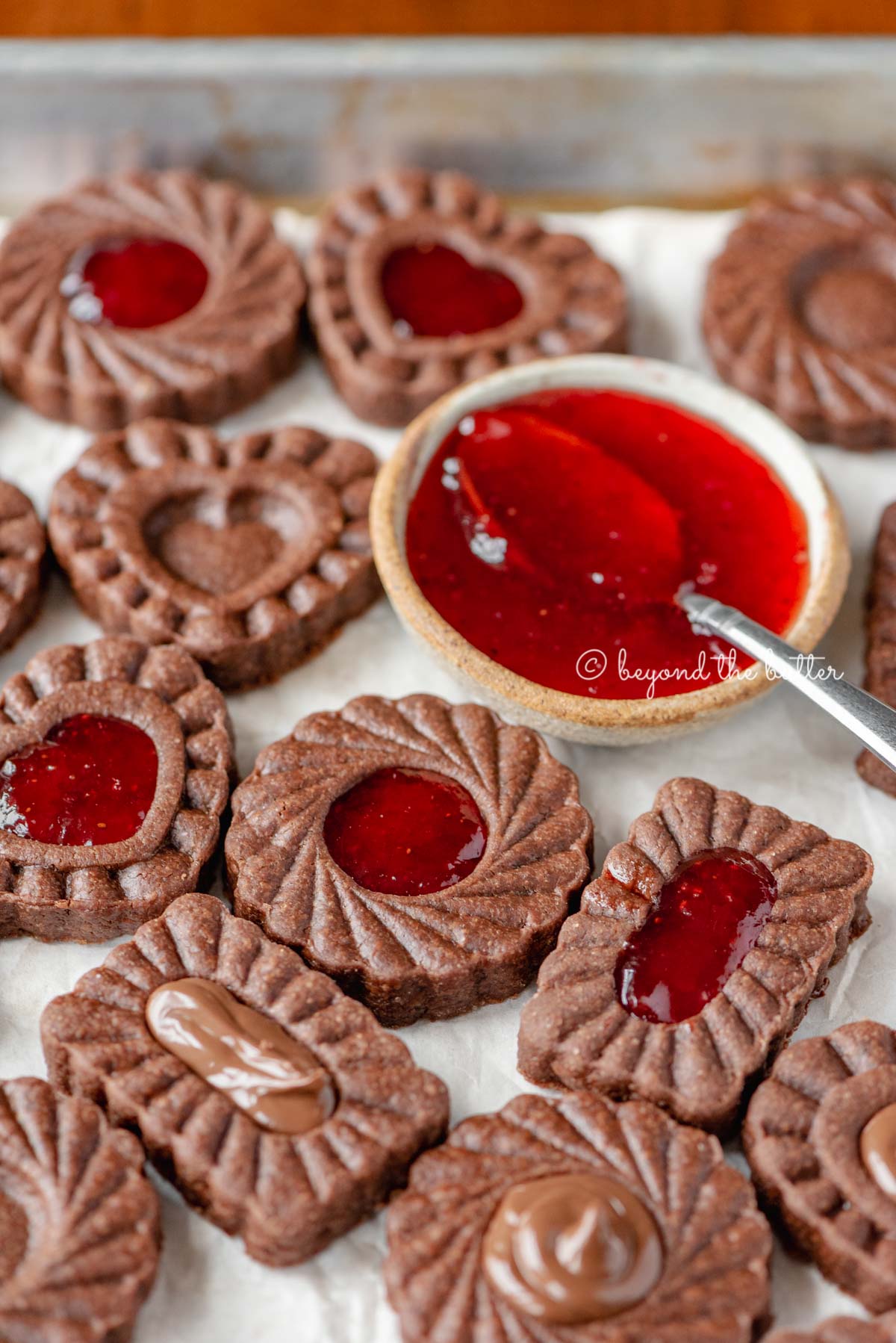 Parchment lined baking sheet full of chocolate thumbprint cookies with small bowl of strawberry jam and spoon | All Images © Beyond the Butter®