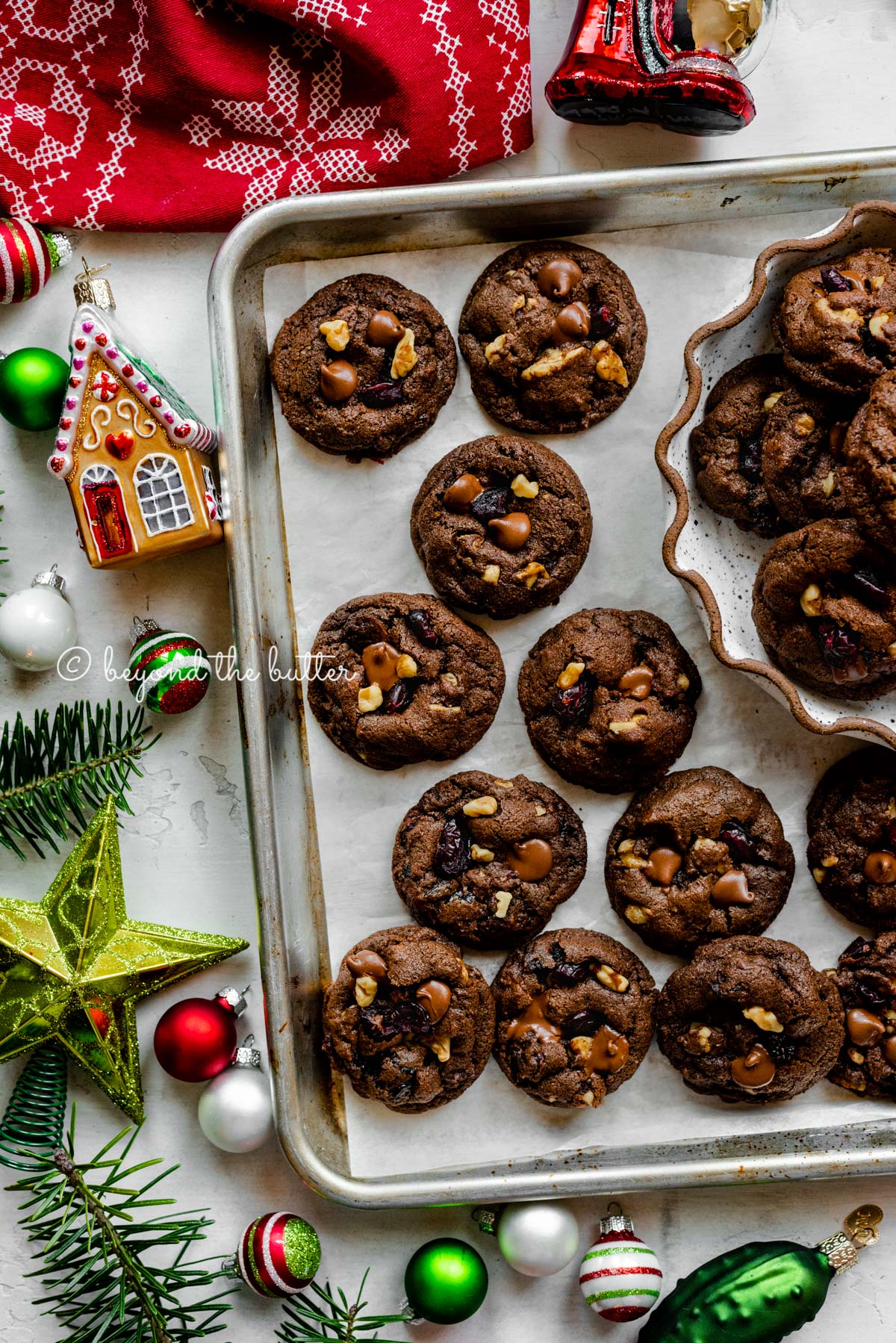 Parchment paper lined baking sheet with dark chocolate cranberry walnut cookies surrounded Christmas tree ornaments | All Images © Beyond the Butter®
