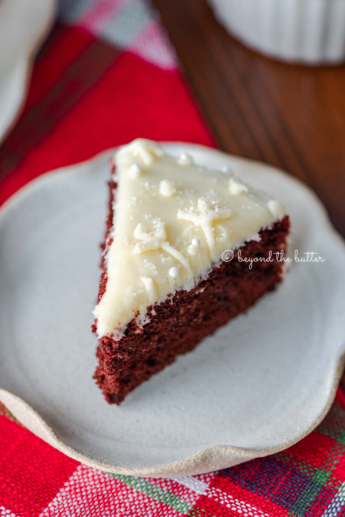 Slice of red velvet cake with cream cheese frosting on a scalloped edge dessert plate with red plaid background | All Images © Beyond the Butter®
