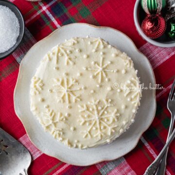Red velvet cake with cream cheese frosting on a scalloped edge dessert plate with red plaid background | All Images © Beyond the Butter®