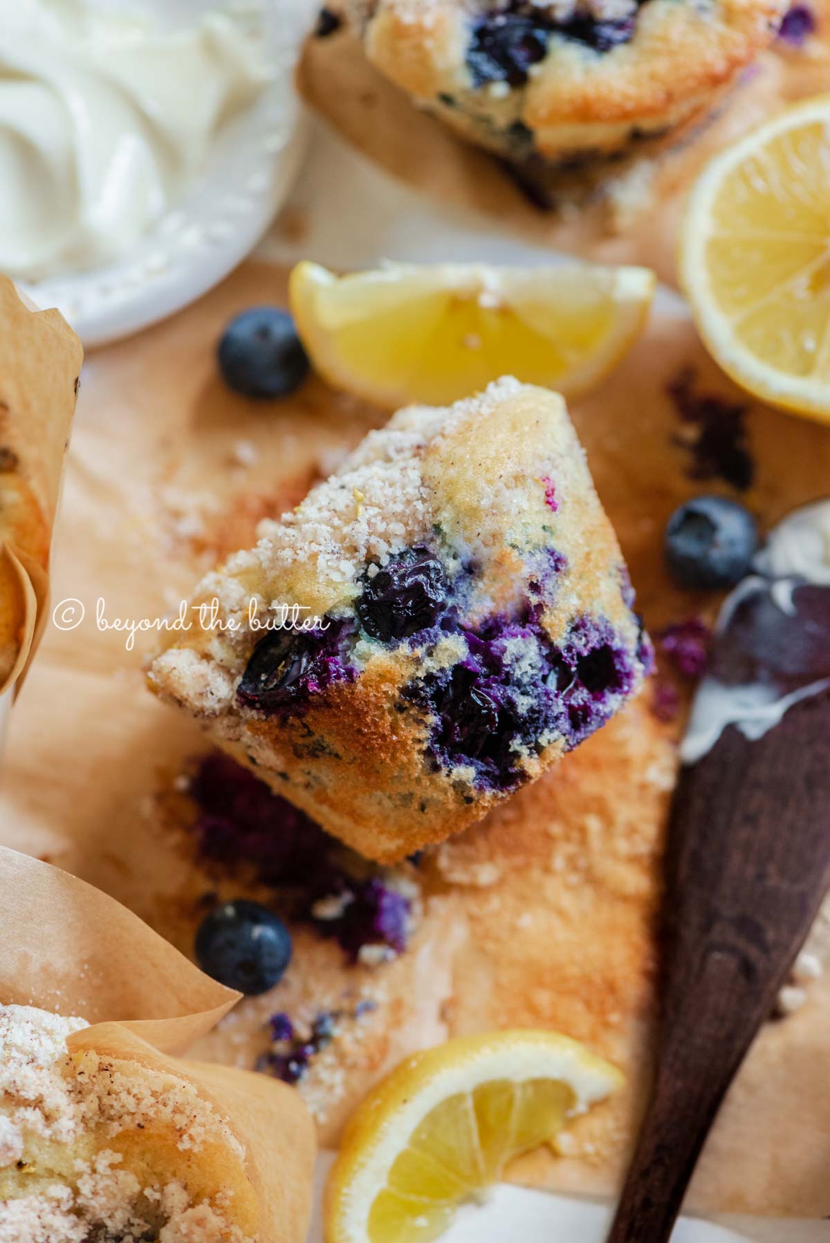Unwrapped lemon blueberry streusel muffin with small bowl of butter, lemon wedges and blueberries around it | All images © Beyond the Butter®