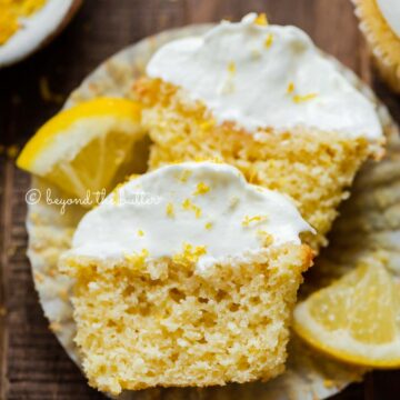 Sliced open lemon cupcake with whipped lemon cream cheese frosting on cupake liner and 2 wedges of lemons on a dark wood background | All Images © Beyond the Butter®