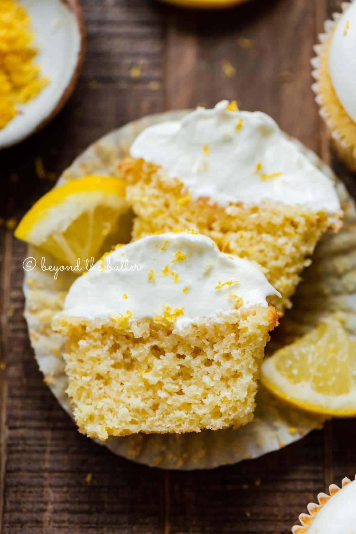 Sliced open lemon cupcake with whipped lemon cream cheese frosting on cupake liner and 2 wedges of lemons on a dark wood background.