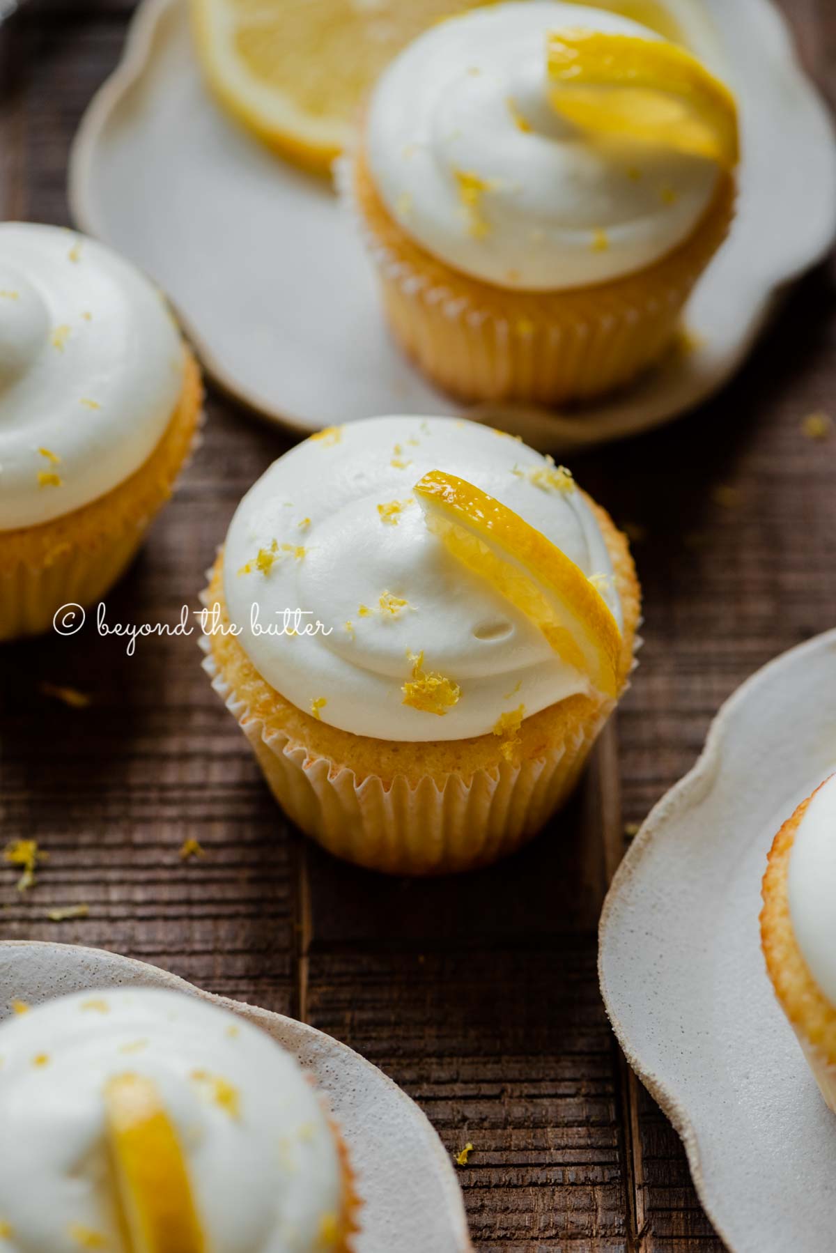 Randomly placed lemon cupcakes with lemon cream cheese frosting, garnished with lemon triangles with a small bowl of lemon zest on a dark wood background | All Images © Beyond the Butter®