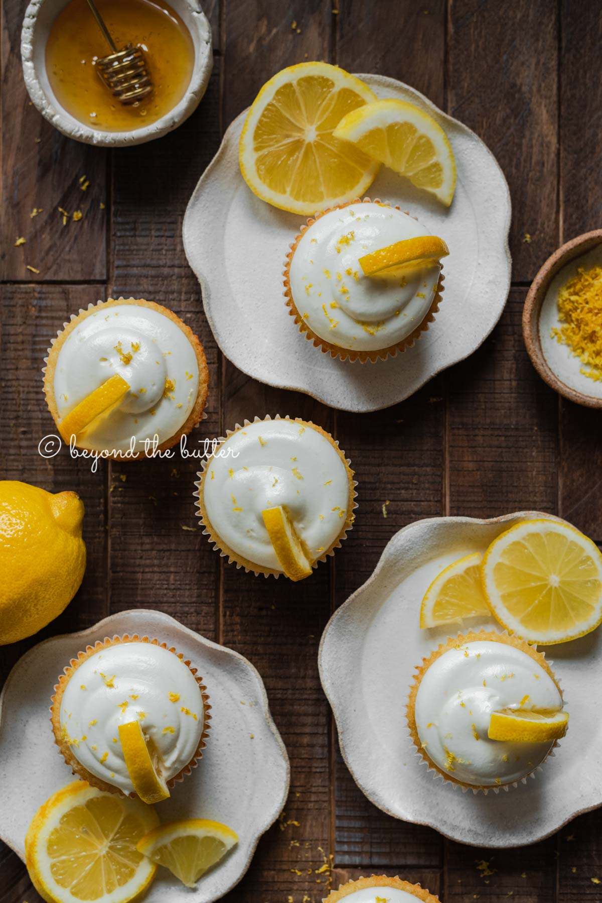 Randomly placed lemon cupcakes with lemon cream cheese frosting, garnished with lemon triangles on a dark wood background | All Images © Beyond the Butter®