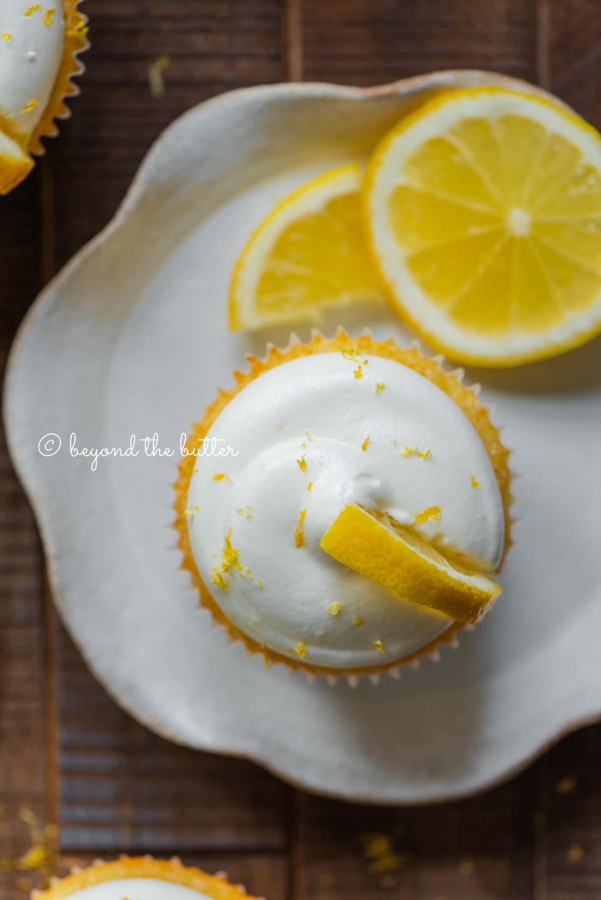 Lemon cupcake with lemon cream cheese frosting garnished with lemons on a small wavy dessert plate | All Images © Beyond the Butter®