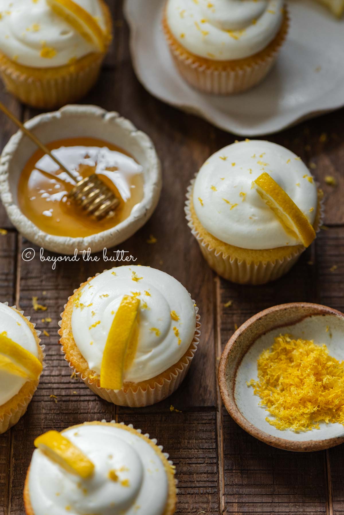 Randomly placed lemon cupcakes with a small bowl of honey and small bowl of lemon zest on a dark wood background | All Images © Beyond the Butter®