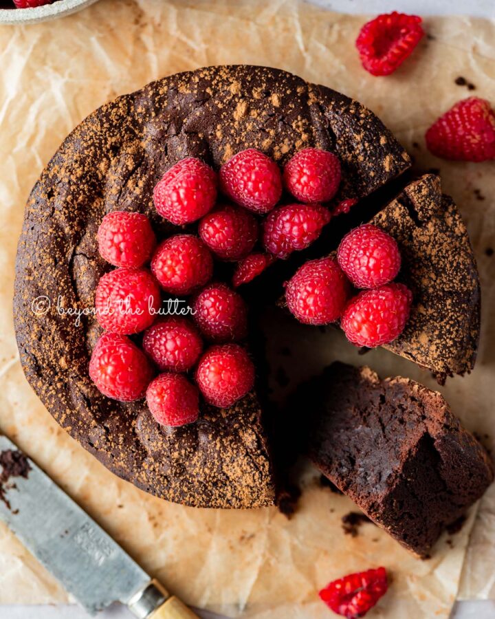 Sliced small flourless chocolate cake topped with cocoa powder and fresh raspberries on parchment paper.