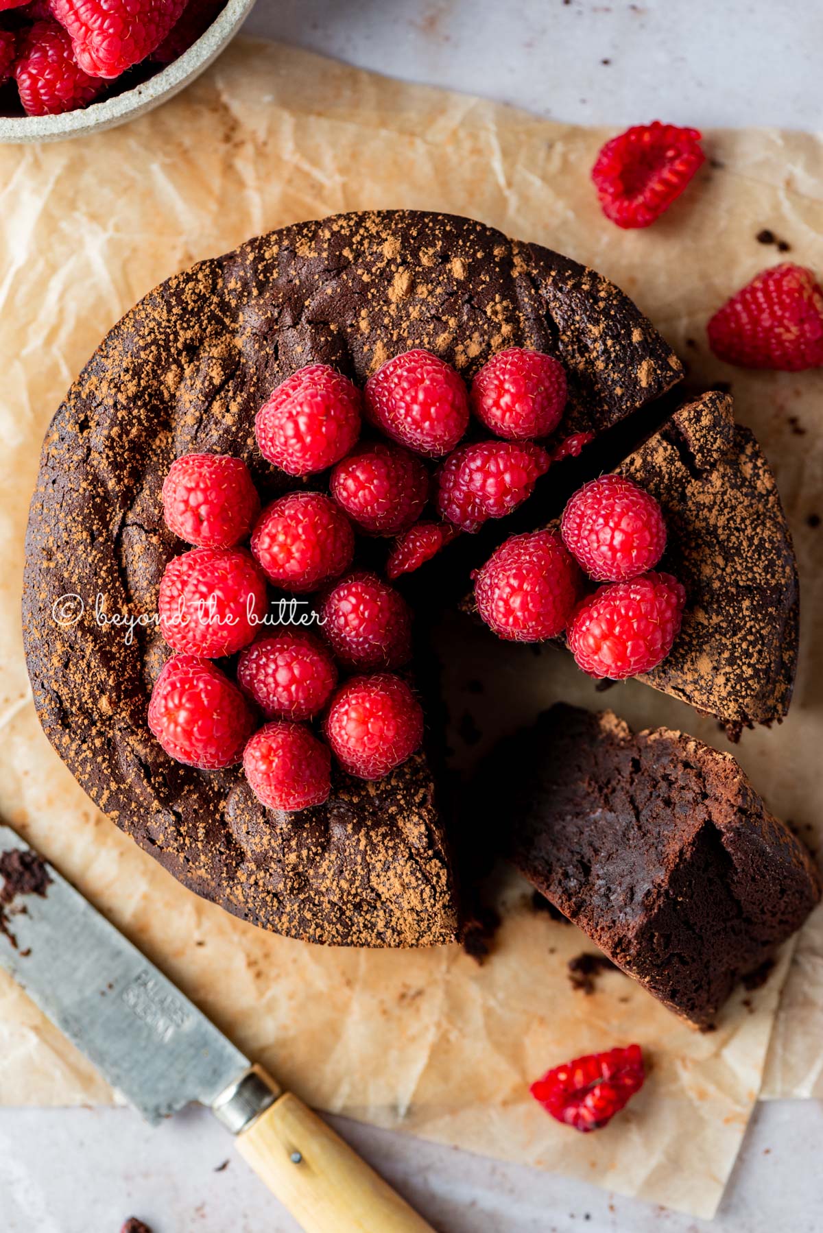 Sliced small flourless chocolate cake topped with cocoa powder and fresh raspberries on parchment paper.
