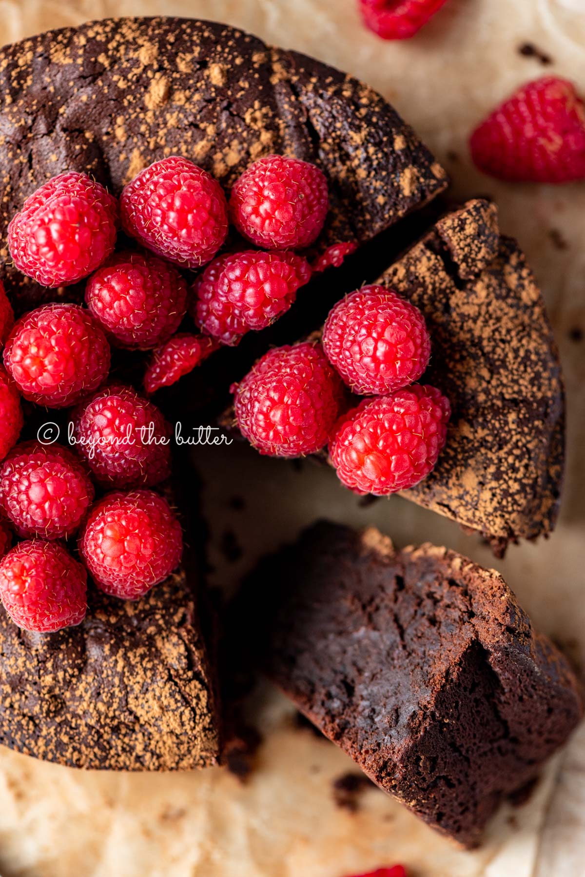 Sliced small flourless chocolate cake topped with cocoa powder and fresh raspberries on parchment paper | All images © Beyond the Butter®