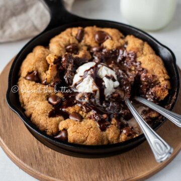 Warm chocolate chip skillet cookie with scoop of vanilla ice cream and two spoons resting in skillet on wood trivet | All Images © Beyond the Butter®