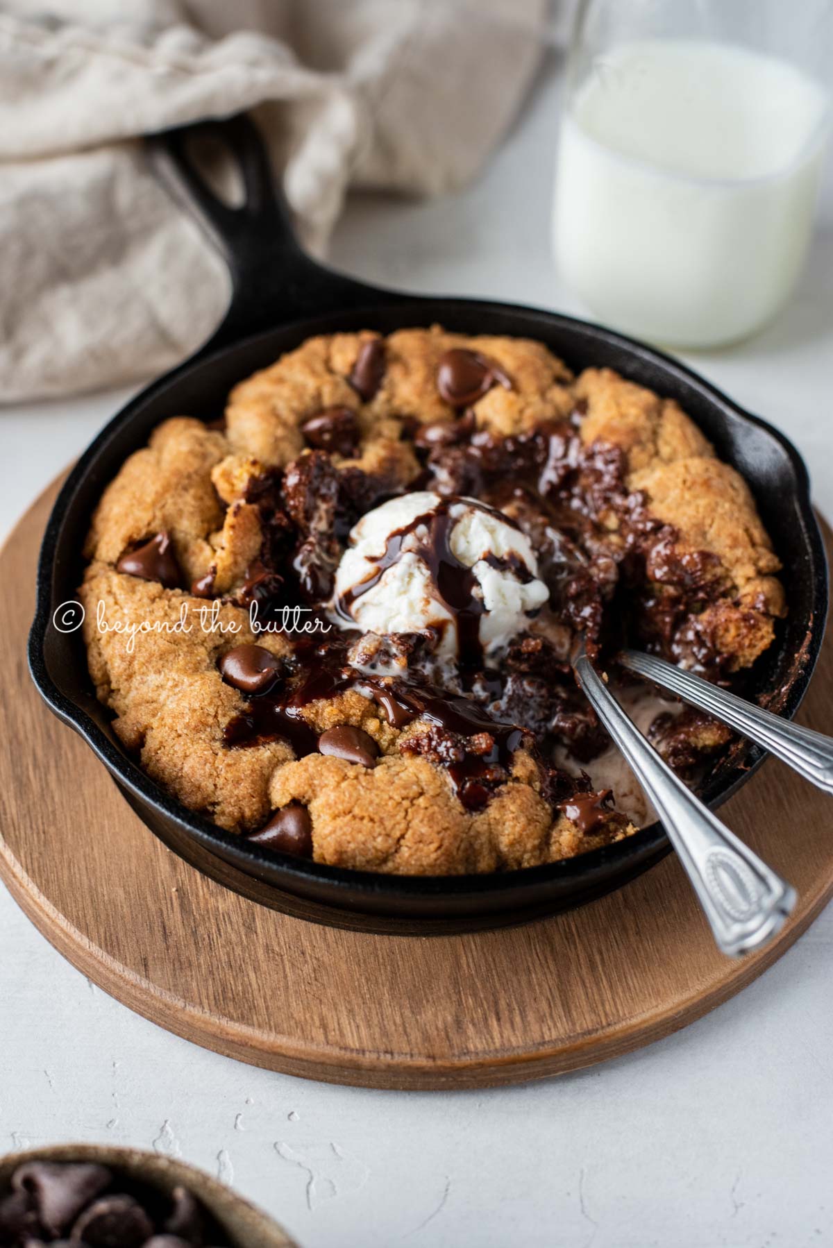 Warm chocolate chip skillet cookie with scoop of vanilla ice cream and two spoons resting in skillet on wood trivet  | All Images © Beyond the Butter®