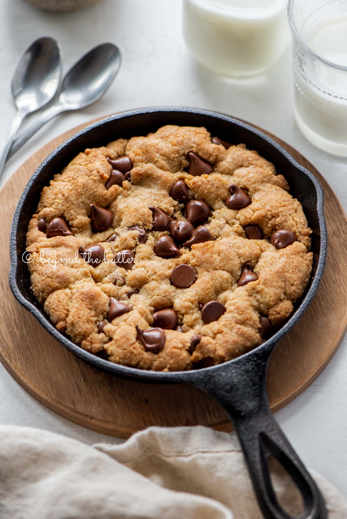 Just baked chocolate chip skillet cookies on light gray background with small bowl of chocolate chips, glasses of milk, and cloth napkin | All Images © Beyond the Butter®